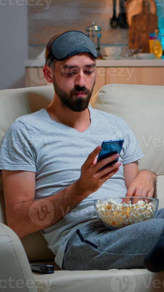 Bored man sitting on couch holding popcorn bowl while texting message on social network using modern phone. Caucasian male watching tv sports series late at night in kitchen photo