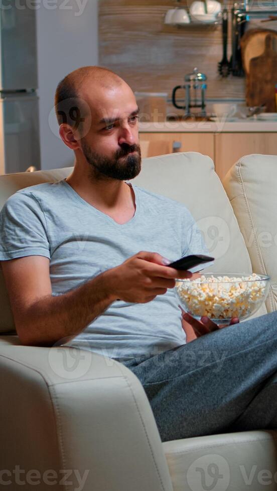 Bored man switching TV channels, holding popcorn in hads late at night while trying to watch entertainment television. Using remote control to choose favourite station and enjoy quality time photo