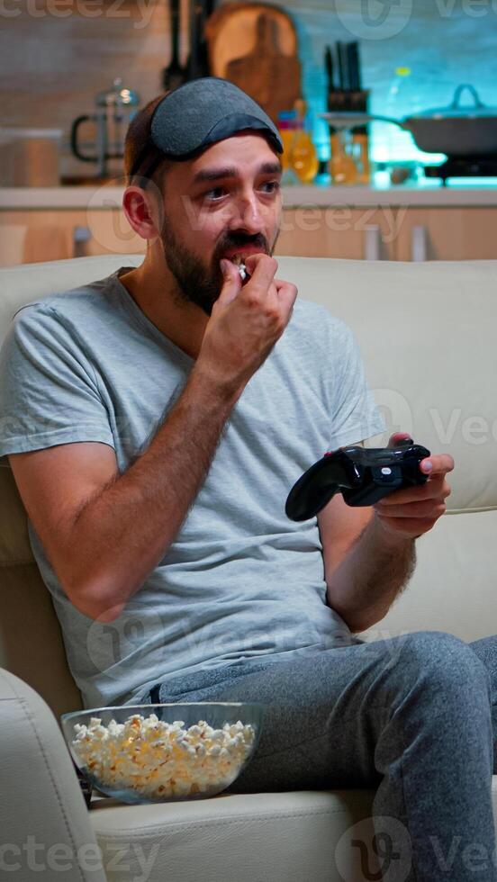 Upset pro gamer sitting on couch and playing soccer videogames for online competition using gaming joystick. Disappointed man looking at television eating popcorn late at night in kitchen photo