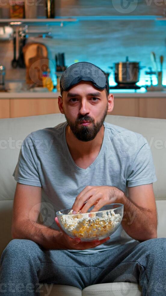 Portrait of man with beard holding popcorn bowl while watching entertainment movie on television. Concentrated person with eye sleep mask sitting on sofa late at night in kitchen photo