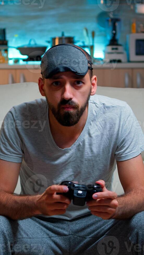 Front view of pro gamer sitting on sofa in front of television while playing videogames competition using wireless joystick. Cheerful man with sleep mask enjoying time alone late at night in kitchen photo