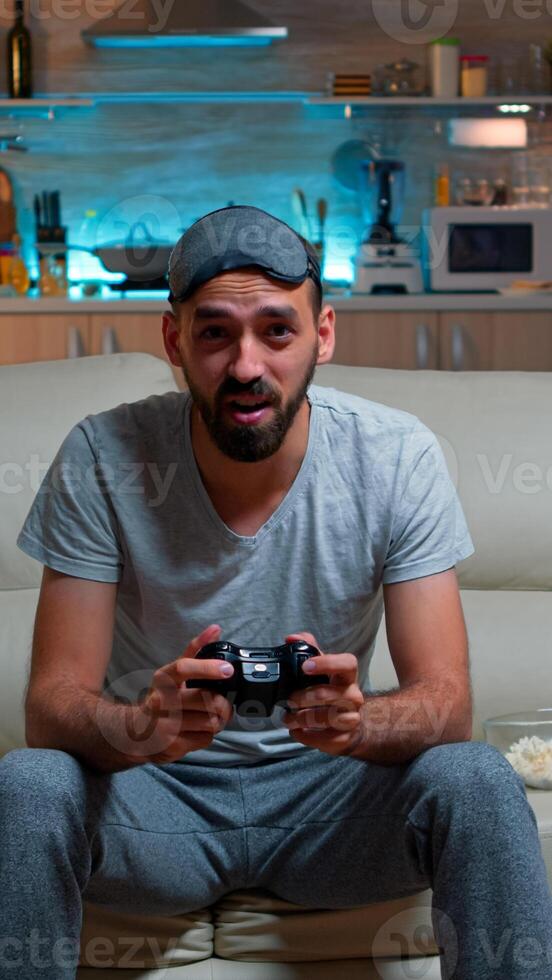Upset pro gamer sitting in front of television losing soccer games holding wireless controller. Frustrated man with sleep mask lying on sofa late at night in kitchen photo