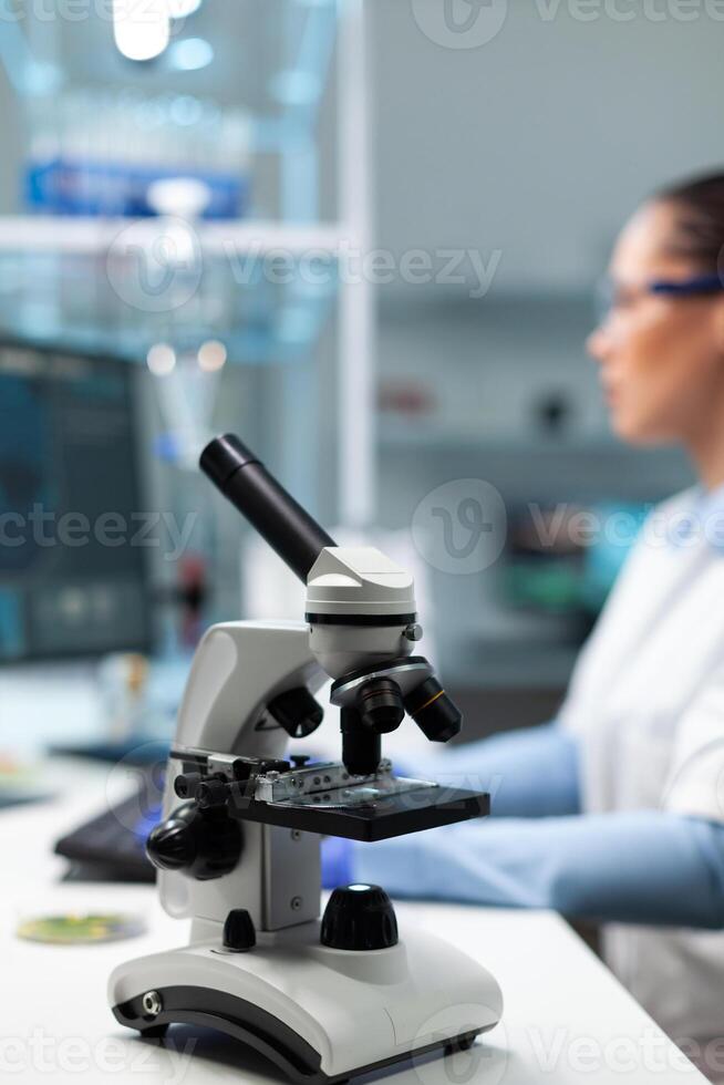 Selectiv focus on medical microscope standing on table in biochemistry hospital laboratory. Specialist microbiologist woman doctor working at biology experiment typing healthcare expertise photo