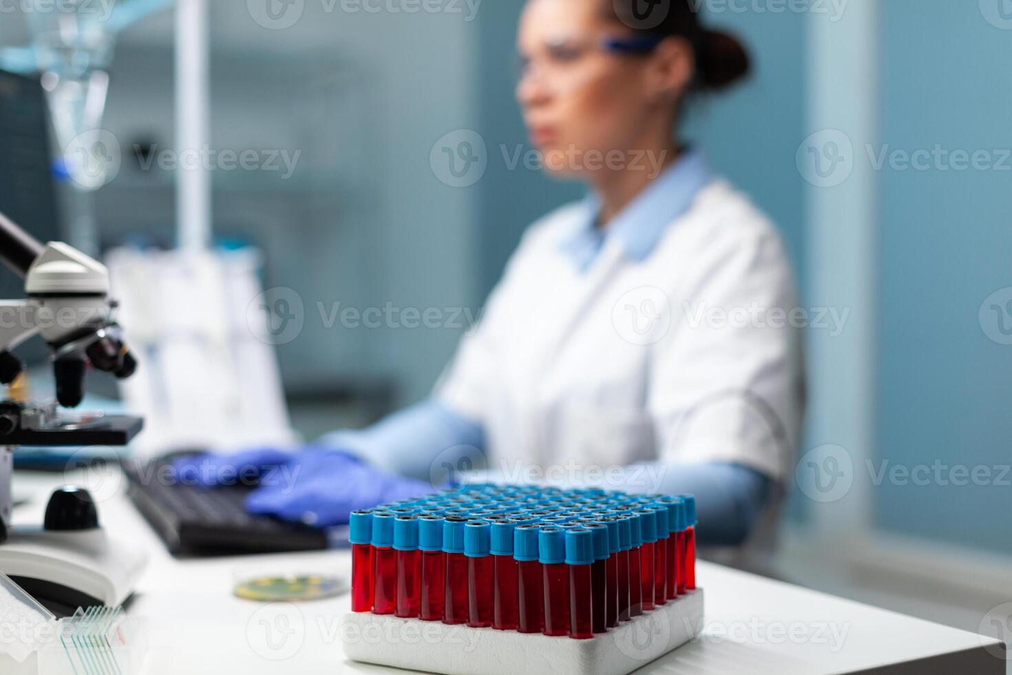 Selective focus on transparent vacutainer with blood standing on table in microbiology hospital laboratory. Scientist biochemist doctor typing medical expertise working at virus vaccine photo