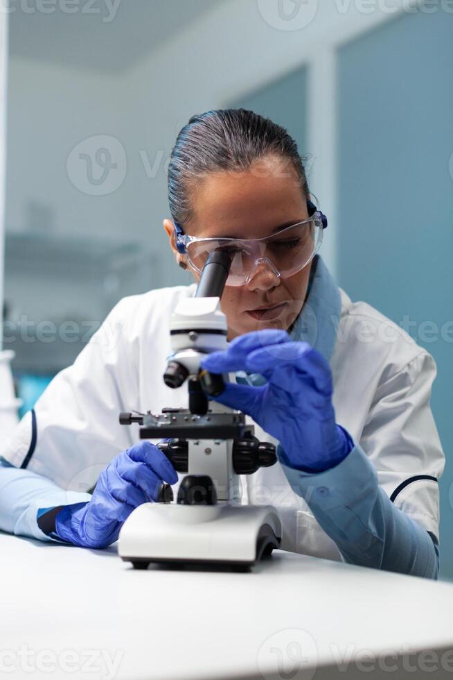 Biologist woman working at vaccine development analyzing blood sample using medical microscope in biochemistry hospital laboratory. Chemist researcher doctor searching for healthcare treatment photo