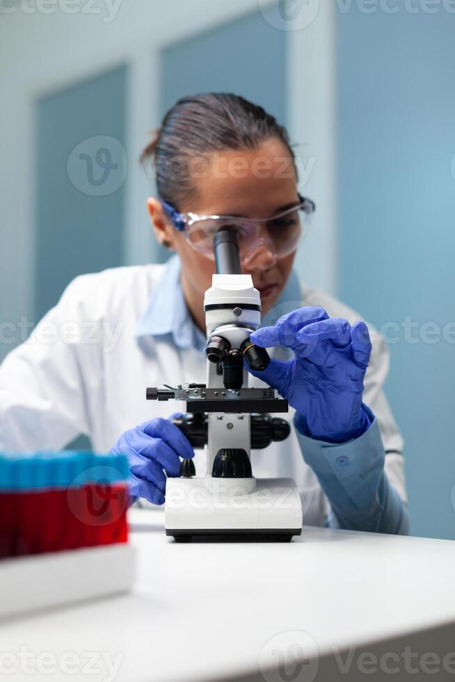 Biologist woman analyzing blood sample using microscope searching for healthcare treatment developing virus vaccine during medical experiment. Chemist researcher working in hospital laboratory photo