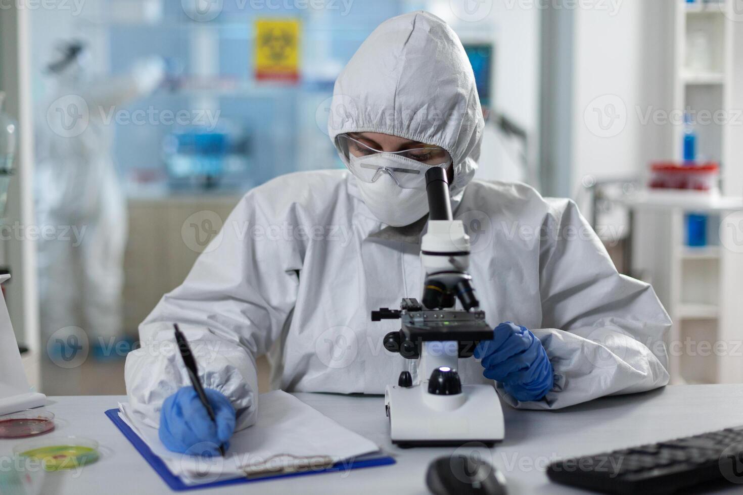 Biologist researcher wearing protective suit analyzing chemicals using medical microscope while writing clinical expertise during scientific experiment. Specialist doctor working in biochemistry lab photo