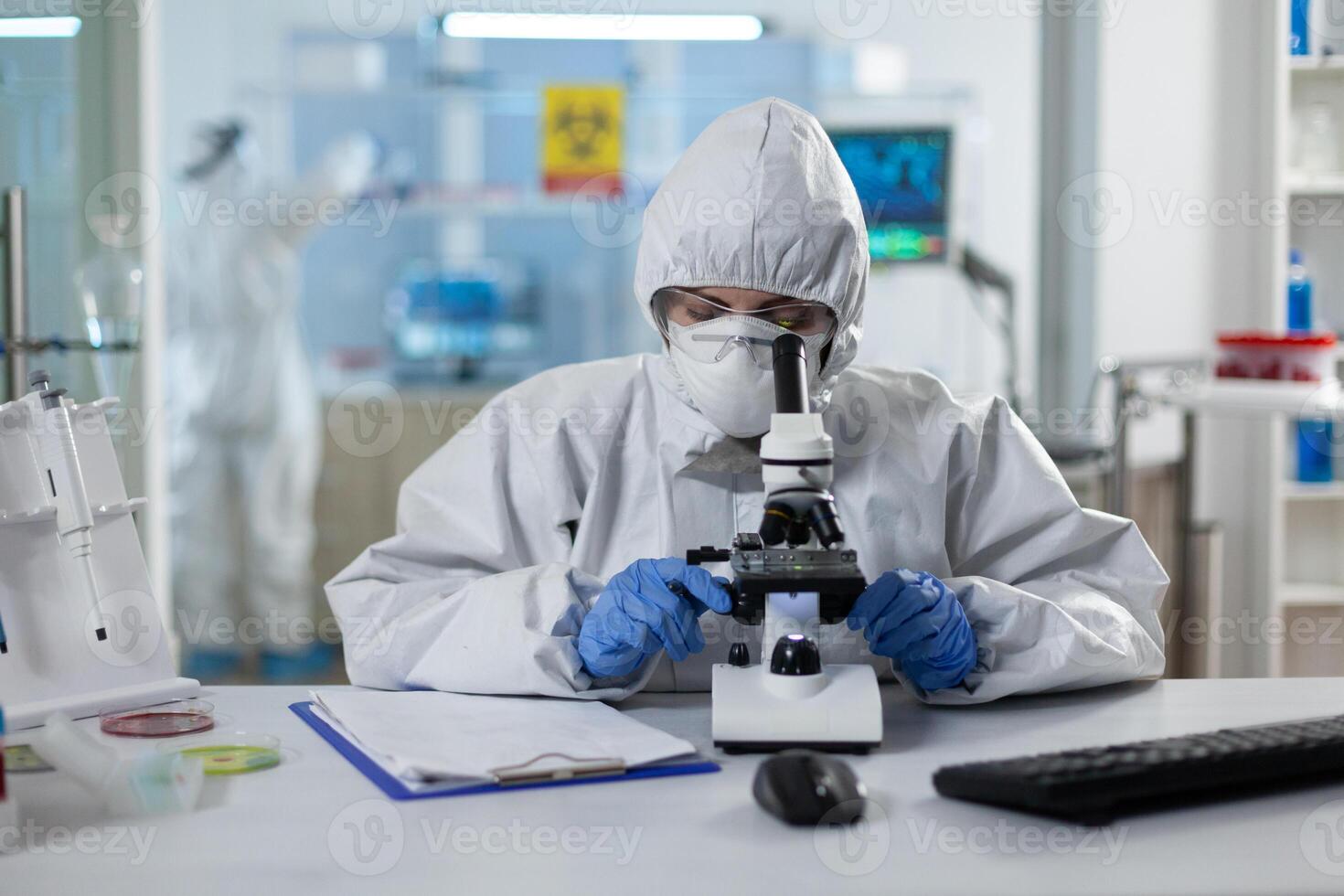 Biologist researcher wearing ppe medical suit analyzing genetics sample using clinical microscope in microbiology hospital laboratory. Scientist woman working at vaccine development expertise photo