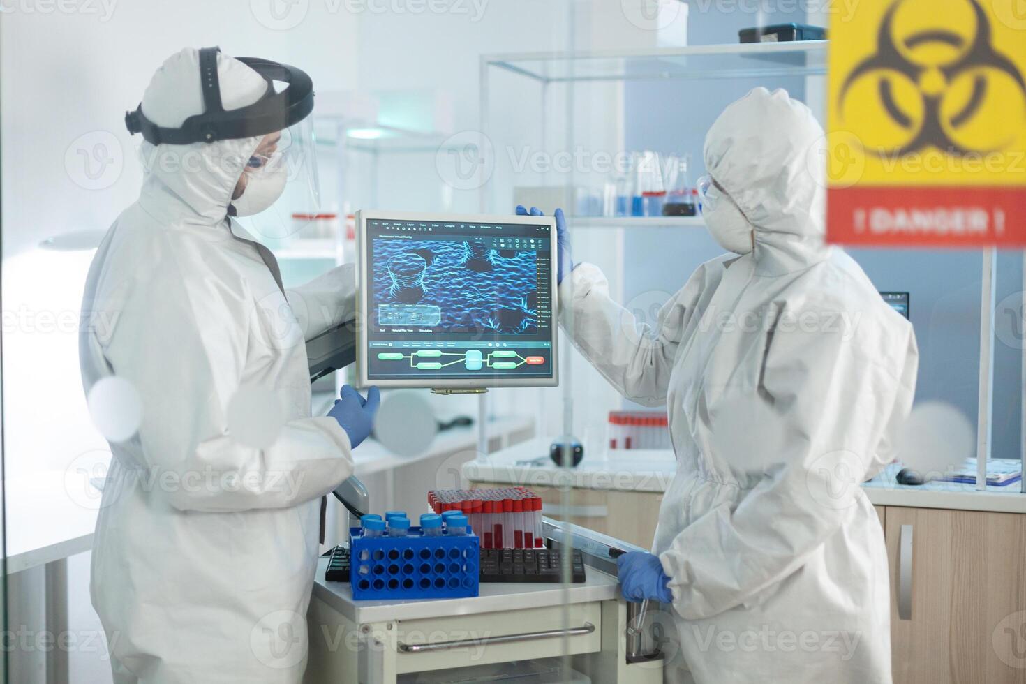 Medical teamwork with protective equipment against covid19 analyzing virus development on computer working at clinical vaccine in microbiological hospital laboratory. Danger sign on glass photo