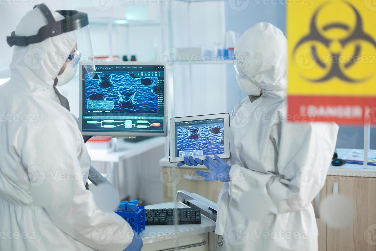 Medical teamwork of scientists wearing protective suit analyzing virus development on tablet during clinical experiment in microbiological hospital laboratory. Danger sign on glass photo