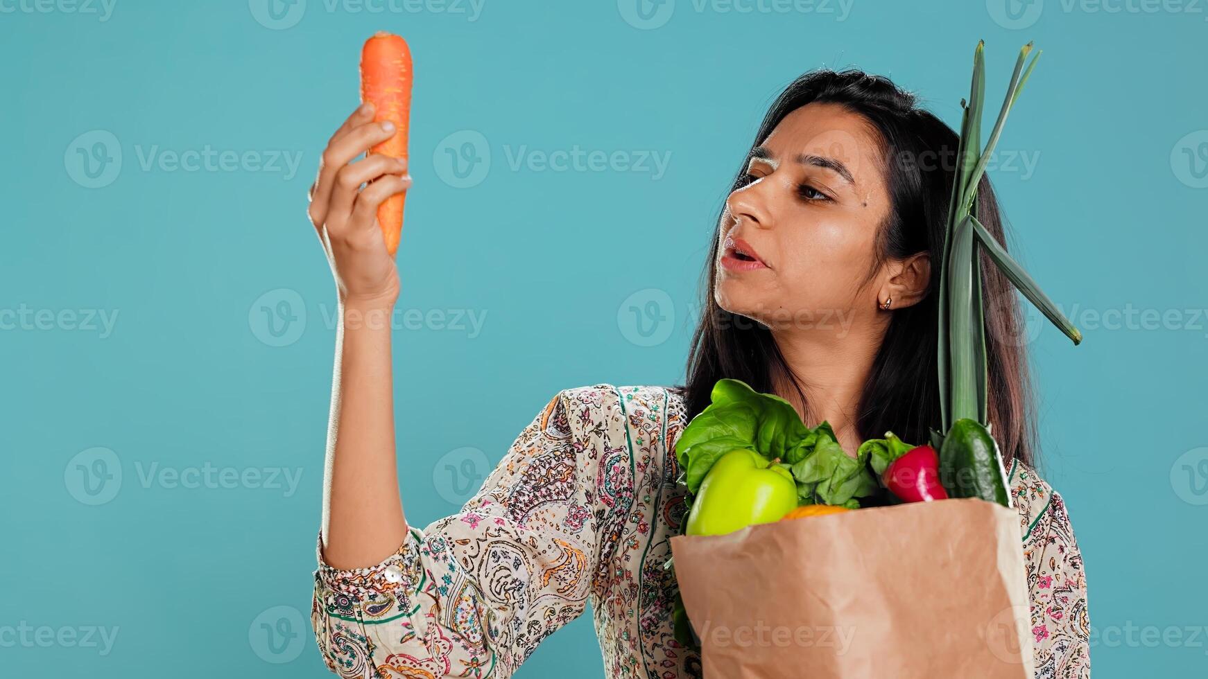 Woman with paper bag in hands filled with vegetables checking carrot for impurities. Conscious living customer with purchased groceries to be used as cooking ingredients, studio background, camera B photo