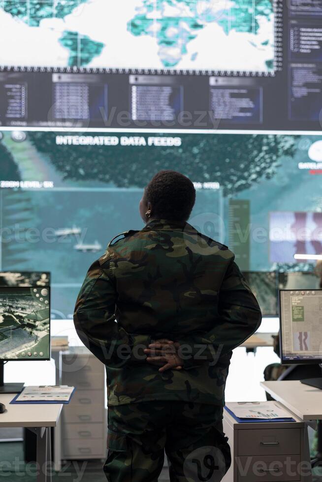 Woman army conscript examines big screen with surveillance imagery in power base monitoring room, analyzing important live transmission to set mission objectives and resources. GPS tracking. photo