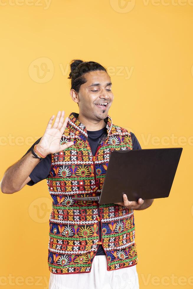 Smiling man doing waving hand gesture, catching up with friend over online videocall on laptop. Upbeat person saluting mate on videoconference using notebook device, studio background photo