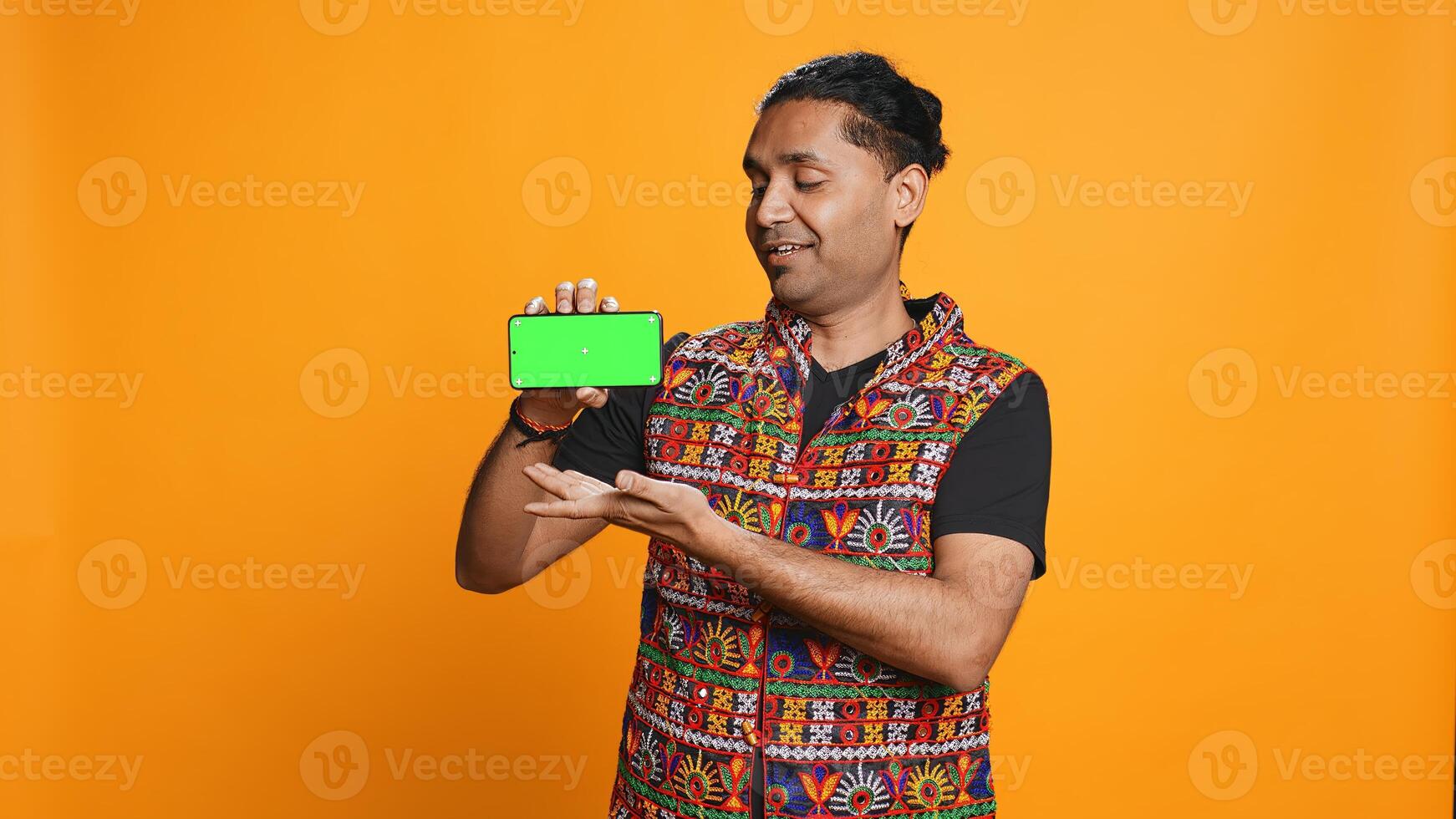 Content creator presenting green screen mobile phone, isolated over studio background. Indian person reviewing copy space mockup smartphone device for internet channel, camera B photo