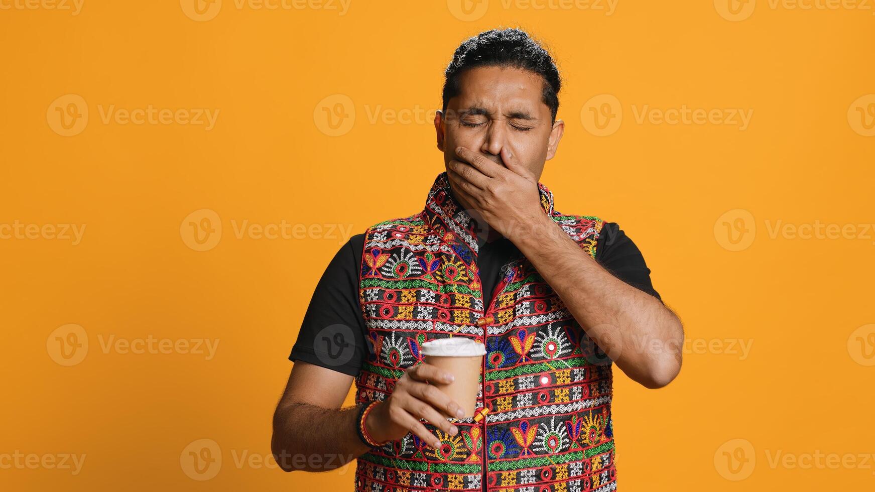 Tired indian man sipping fresh coffee from disposable paper cup early in the morning to wake up and be energized. Person drinking hot beverage from recycled takeaway cup, studio background, camera A photo
