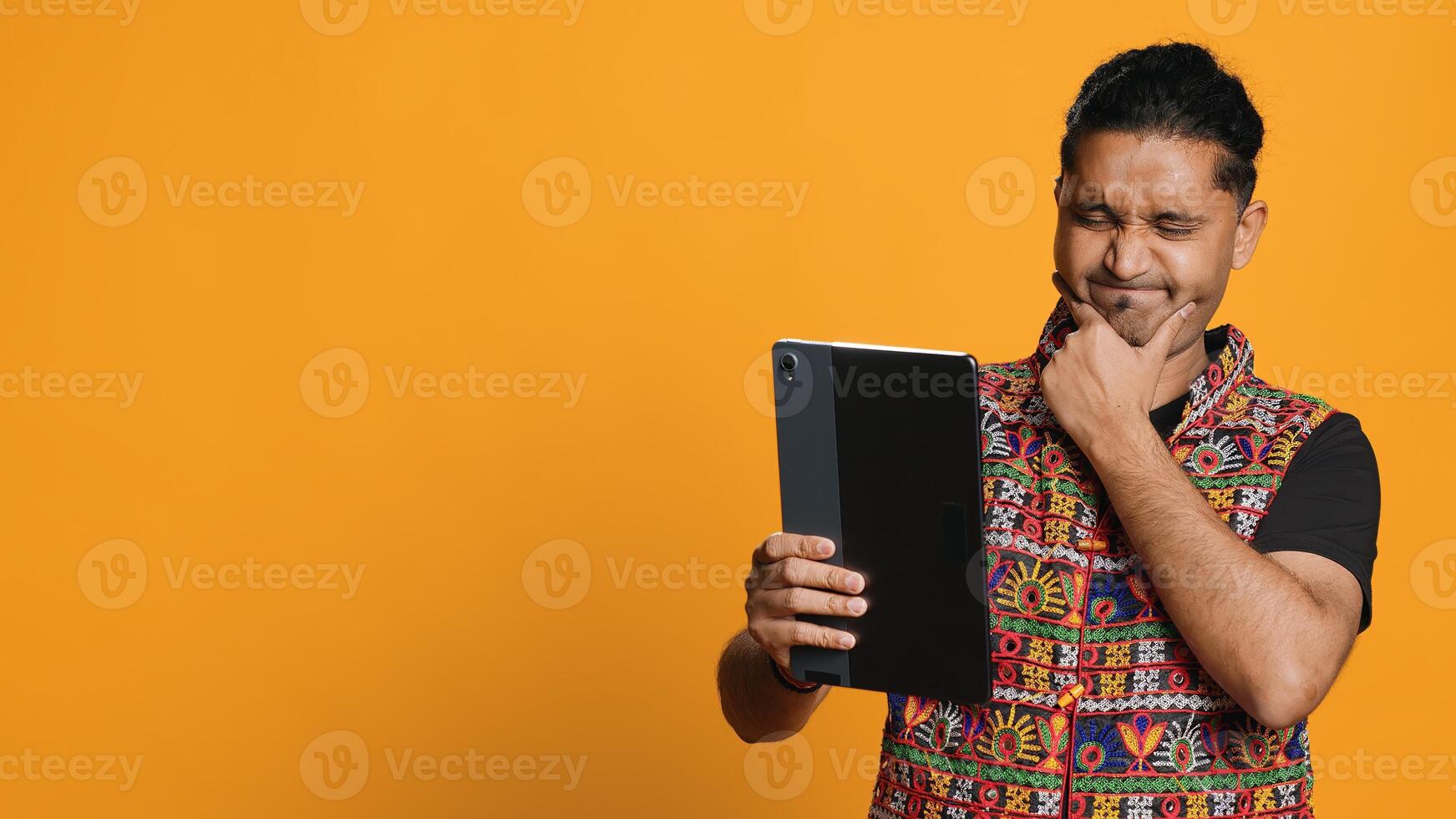 Relaxed man having entertaining conversation with friend over online videocall on device. Carefree indian person discussing with mate on teleconference using tablet, studio backdrop, camera A photo