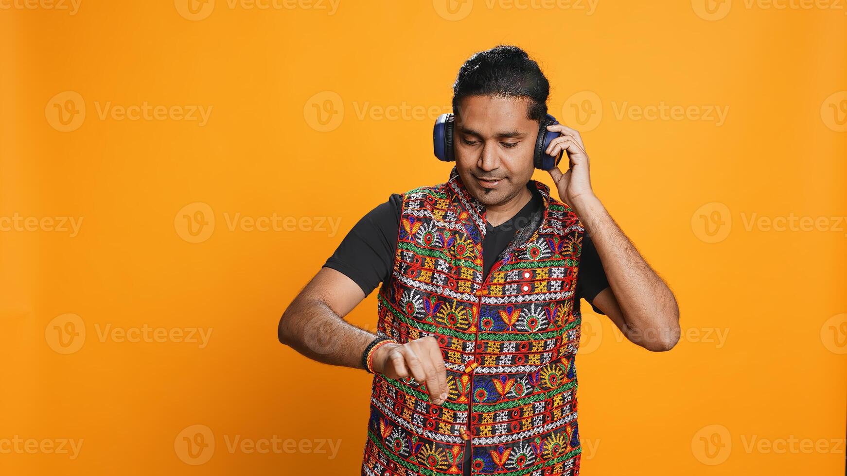 Indian man playing music, having fun moving on rhythm, pretending to use turntables. Upbeat DJ wearing headphones, producing beats, isolated over studio background, camera B photo
