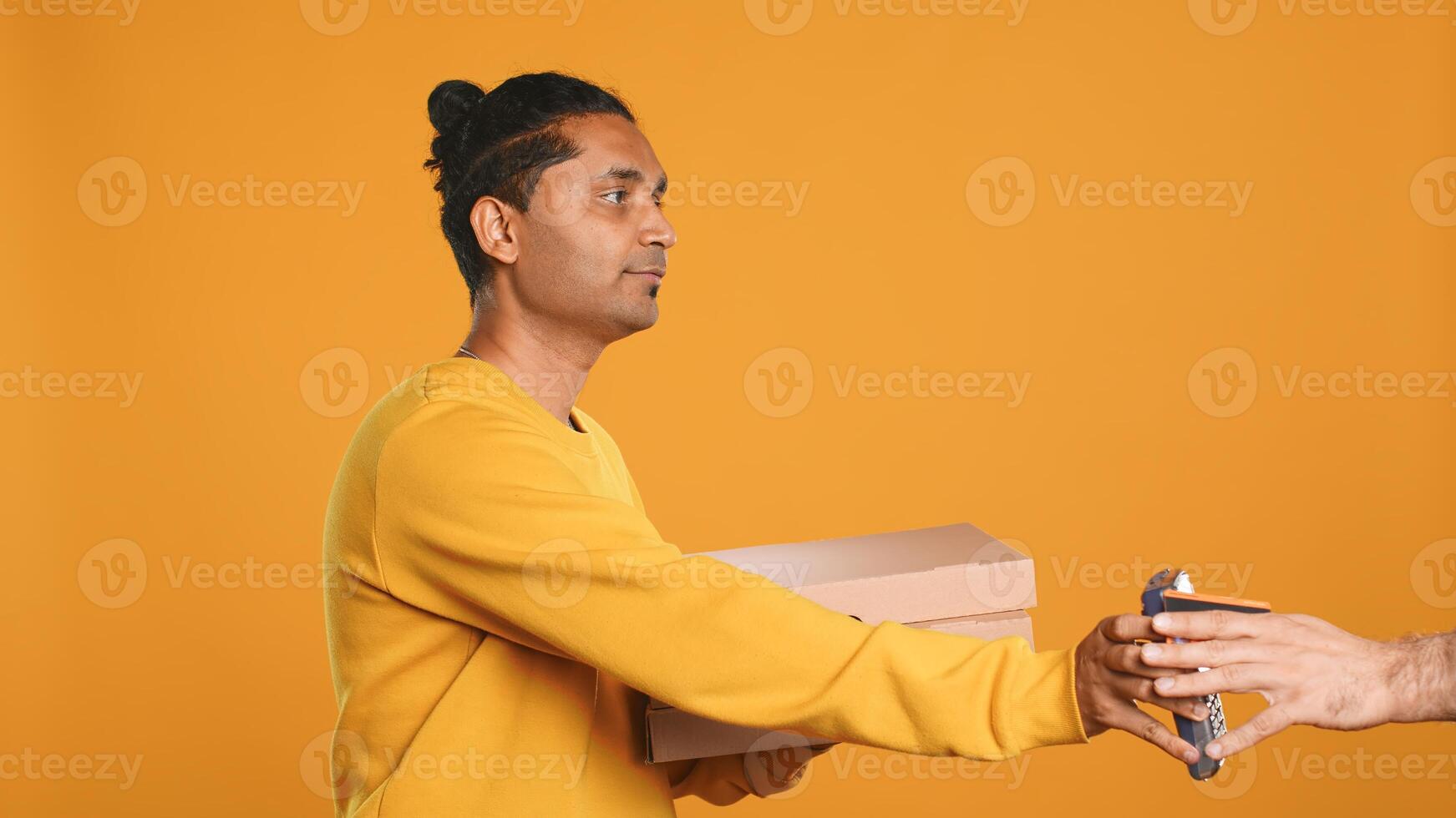Indian delivery worker offering takeaway order to customer paying with debit card. Client buying meal from courier, swiping card on POS device, studio background, camera A photo