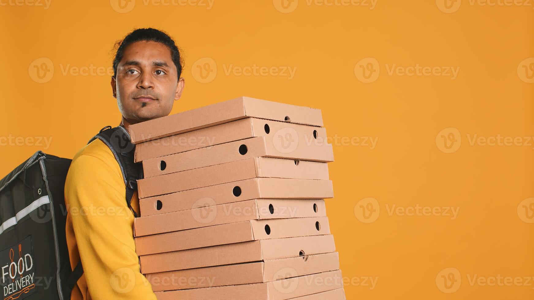 Indian pizza delivery worker carrying takeaway orders, wearing thermal backpack. Courier holding lunch boxes and transportation bag, rushing to clients, isolated over studio background photo