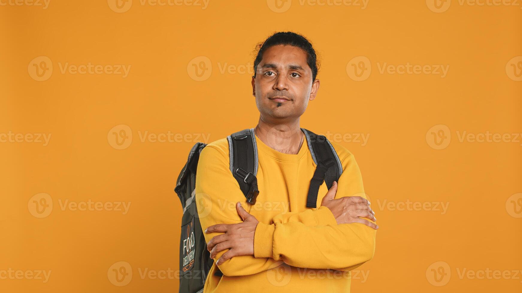 Portrait of confident indian man holding thermal backpack, crossing arms, studio background. Radiant nonchalant delivery person prepared to deliver takeaway food to customers, camera A photo