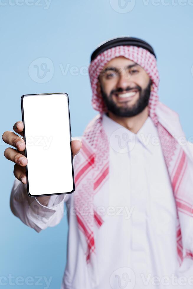 Smiling muslim man presenting smartphone empty screen with copy space for software app promotion. Cheerful person in arabic clothes showing mobile phone with blank display closeup photo