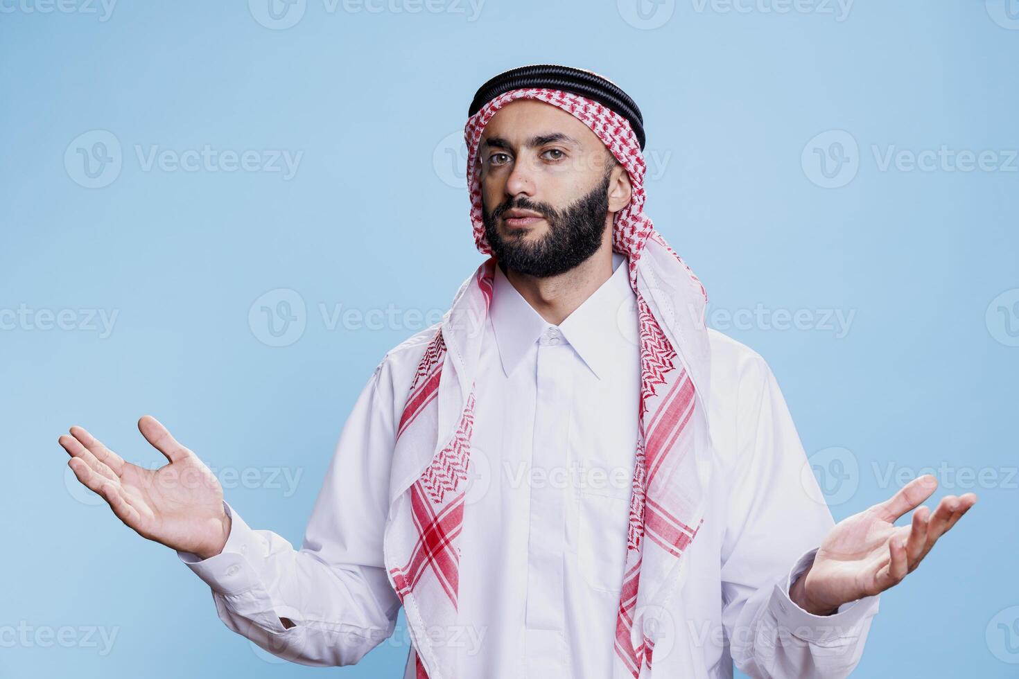 Confused muslim man in traditional attire spreading hands and expressing uncertainty portrait. Puzzled arab person shrugging shoulders with open arms and looking at camera photo