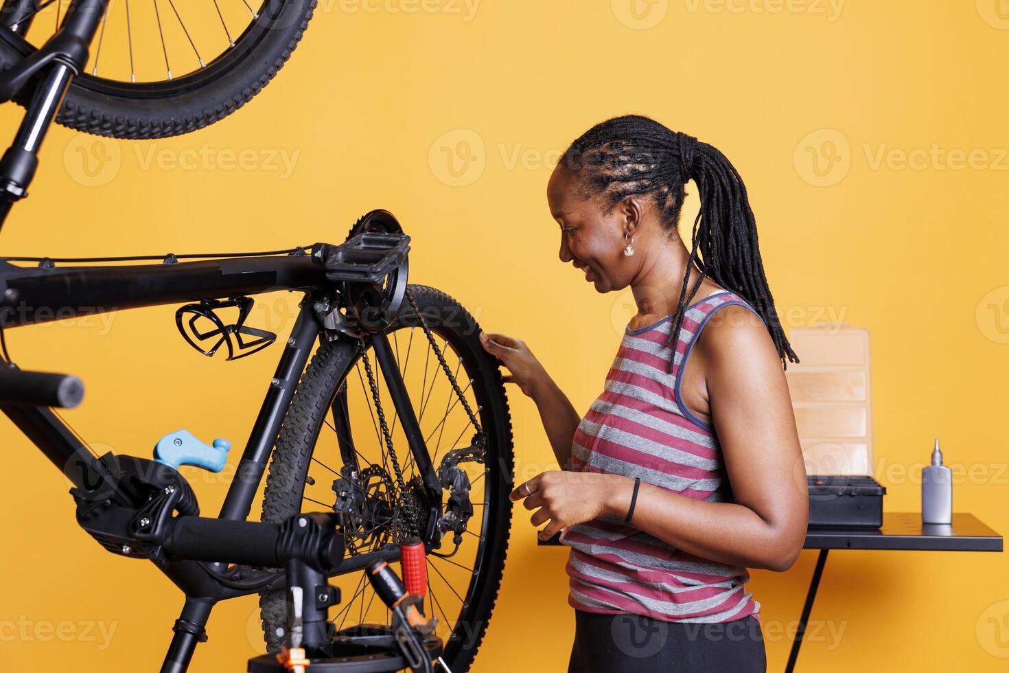 Active young woman does yearly maintenance and adjustment of bicycle tire in front of isolated yellow background. Athletic african american lady examining her bike wheel for recreational cycling. photo