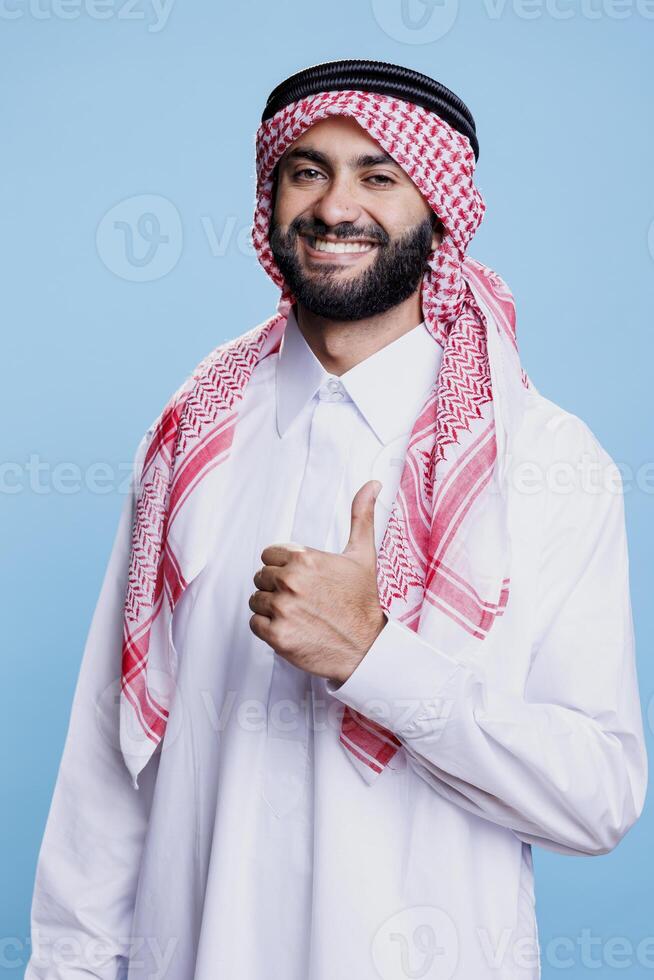Smiling arab man in traditional robe and headdress showing thumb up studio portrait. Muslim person dressed in islamic clothes posing with positive gesture and looking at camera photo