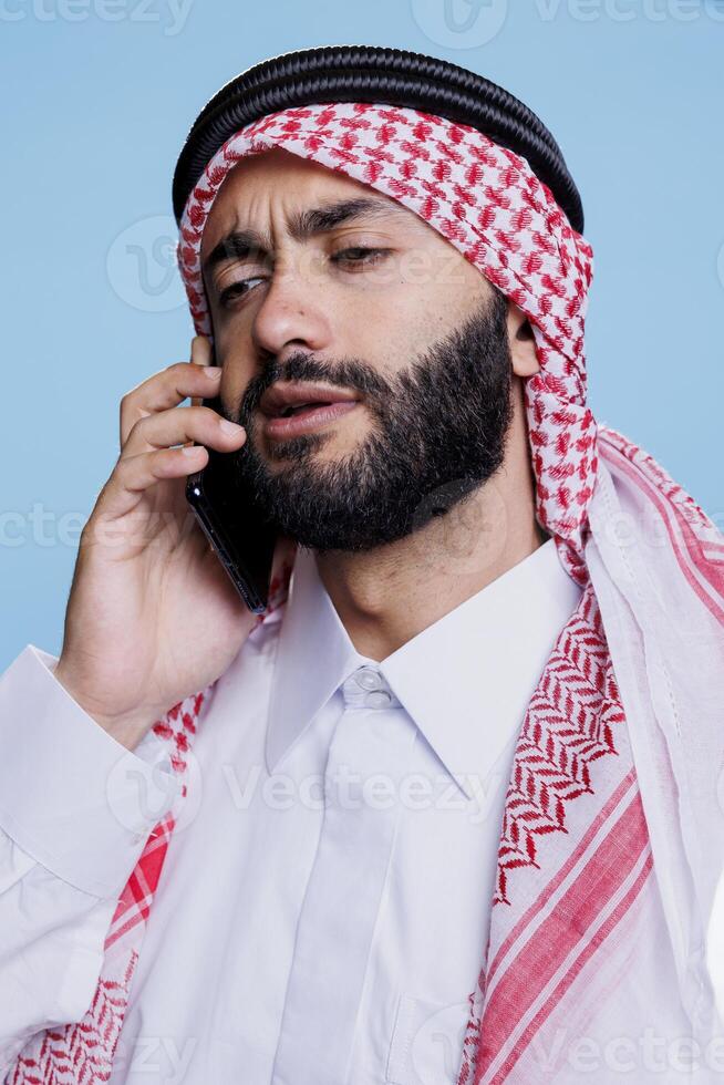 Muslim man wearing traditional headscarf speaking on smartphone with confused expression. Young arab person dressed in red and white checkered ghutra speaking on mobile phone photo