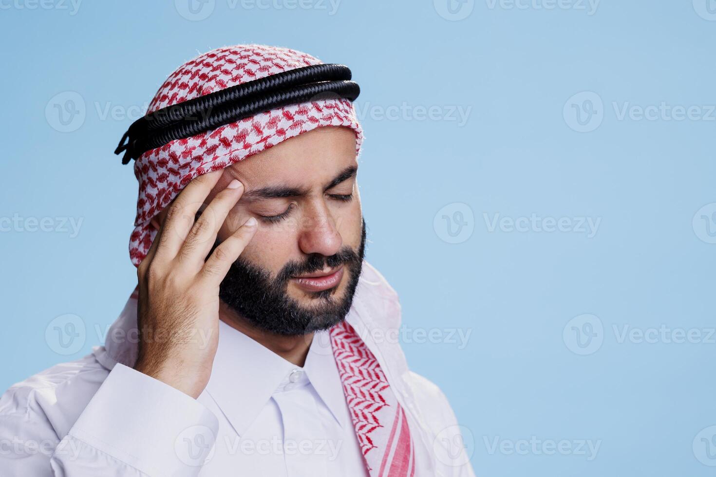 Exhausted muslim man with closed eyes holding temple while suffering from migraine. Arab person wearing traditional clothes feeling pain and touching head while having high blood pressure photo