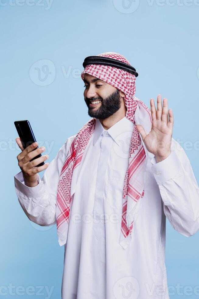 Muslim man greeting with friendly hand wave and smile while chatting in videocall on smartphone. Cheerful arab in thobe saying hello while speaking in online meeting using mobile phone photo