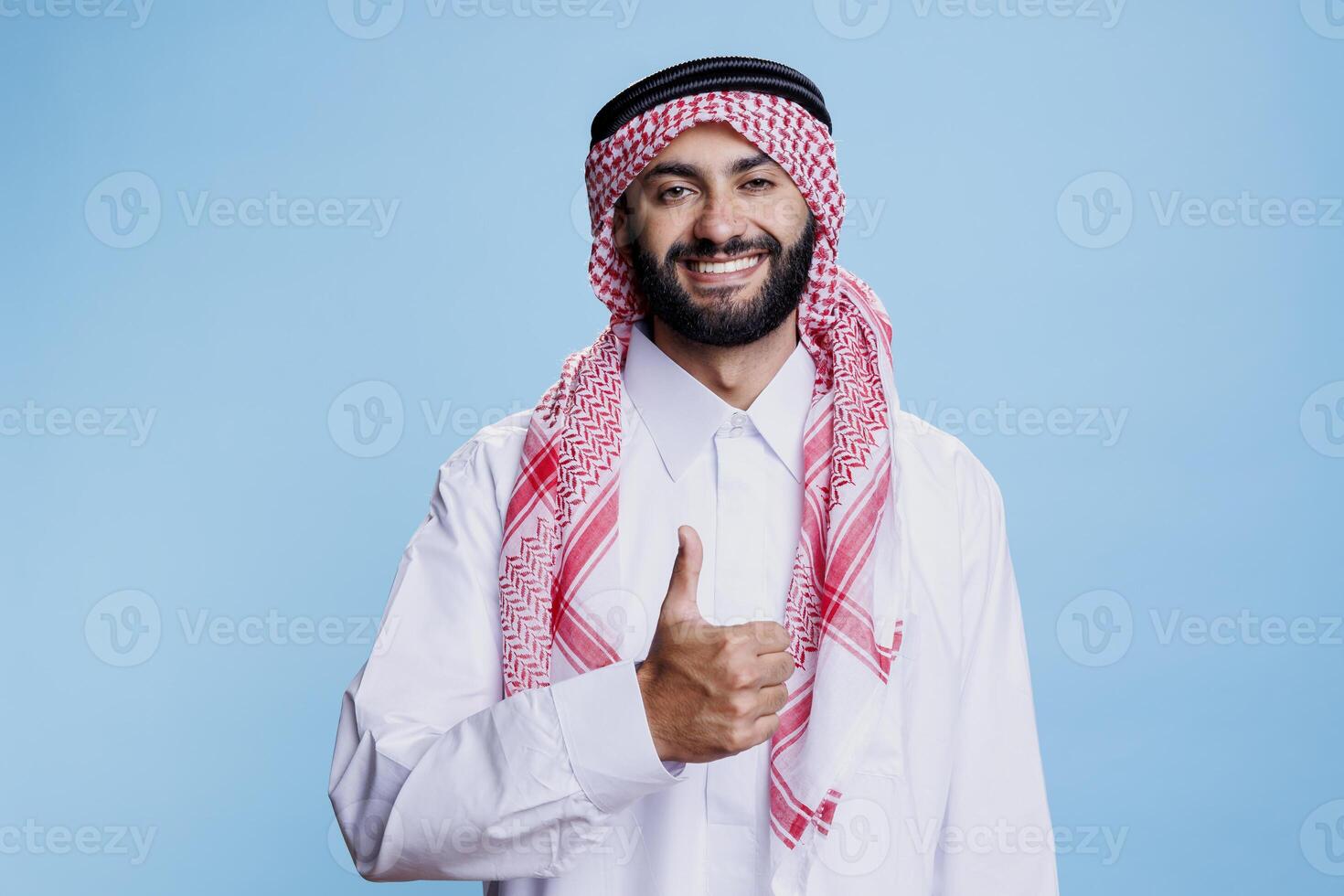 Smiling muslim man in white thobe and checkered headdress showing thumb up studio portrait. Cheerful arab posing with positivity and satisfaction gesture and looking at camera photo