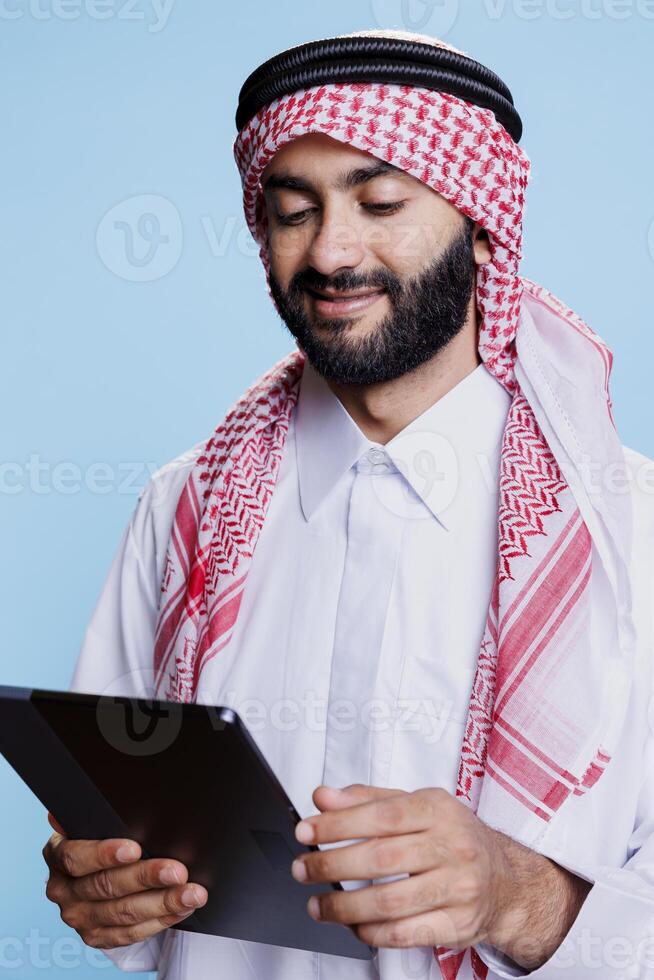 Muslim man wearing traditional clothes holding digital tablet and exploring content. Smiling arab dressed in islamic white thobe and ghutra headscarf scrolling social media on touchscreen photo