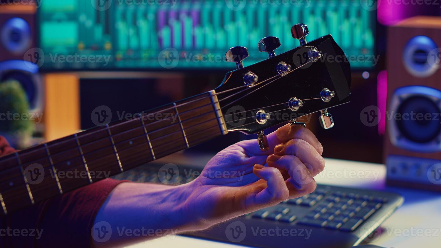 Musical performer tuning his guitar by twisting the knobs, preparing to play acoustic instrument in home studio. Artist recording sounds to create new track, using equalizer mixing gear. Camera A. photo