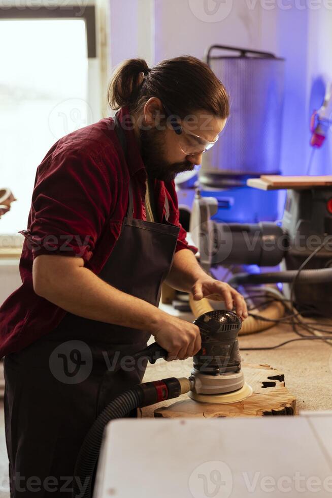Carpenter wearing safety glasses, using orbital sander with finer sandpaper grits to achieve refined finish. Manufacturer using angle grinder on wood block, achieving professional quality results photo