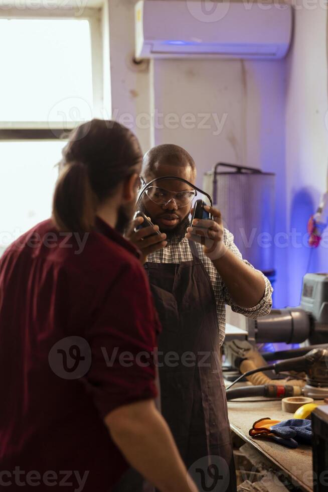 Master in carpentry shop instructing BIPOC worker to wear protection gear, preventing workplace accidents. Cabinetmaker making sure apprentice uses safety earmuffs and goggles to avoid injury photo