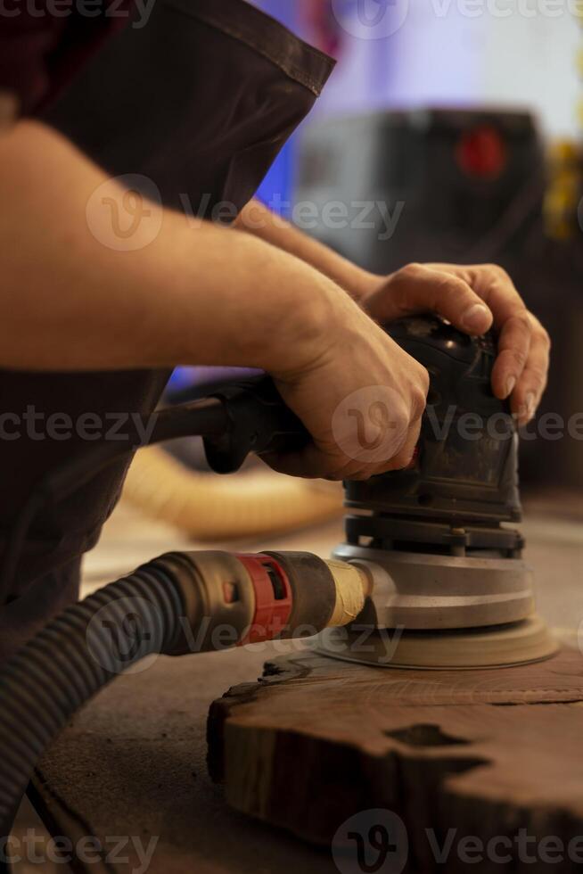 Creative craftsperson using angle grinder on lumber block to smooth surfaces by abrasion with sandpaper. Man using orbital sander gear for furniture assembling work in joinery, close up shot photo