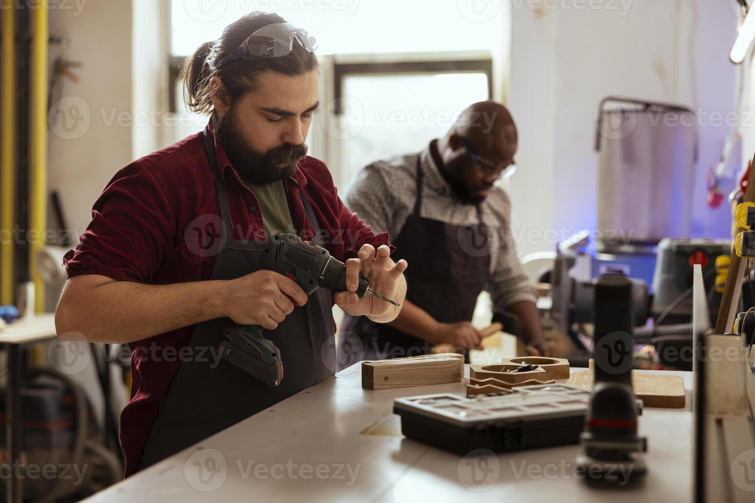 Woodworker next to african american coworker changing power drill head. Carpenter working with BIPOC colleague, preparing to insert screws into wooden surfaces with electric tool photo