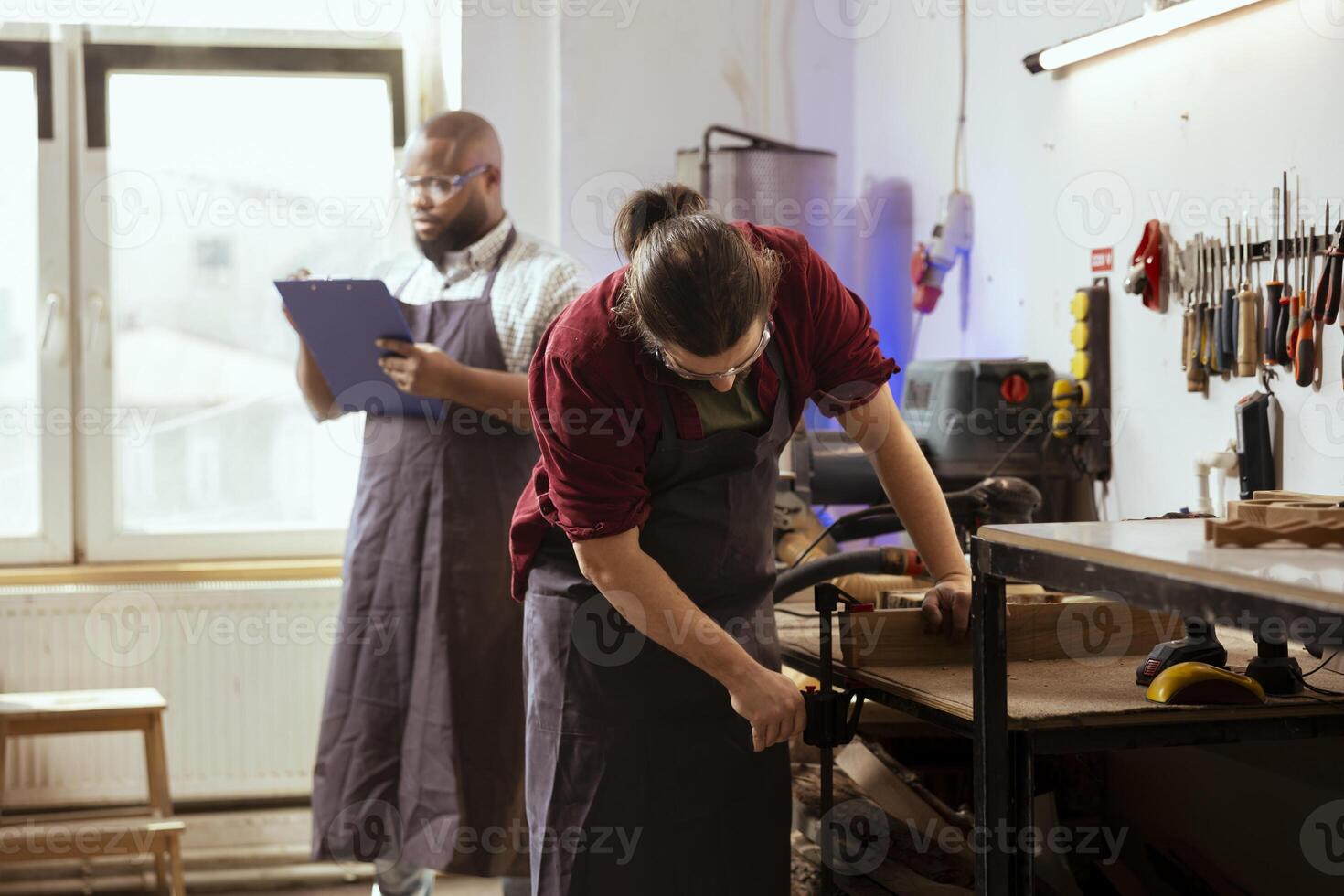 Woodworker wearing safety glasses while using bench vise to hold lumber block in workshop next to coworker. Craftsperson using protective equipment and vice tool to clamp piece of wood photo
