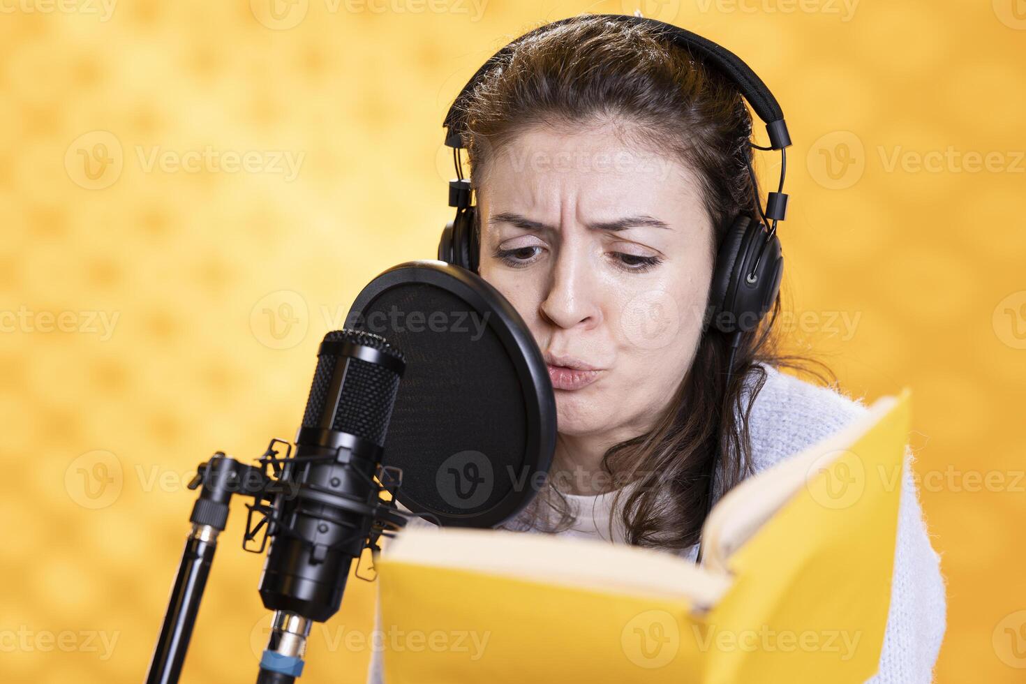 Voice actor with frown on face reading book, recording audiobook using microphone and headset, studio background. Woman using mic to produce digital recording of novel, glowering for dramatic effect photo