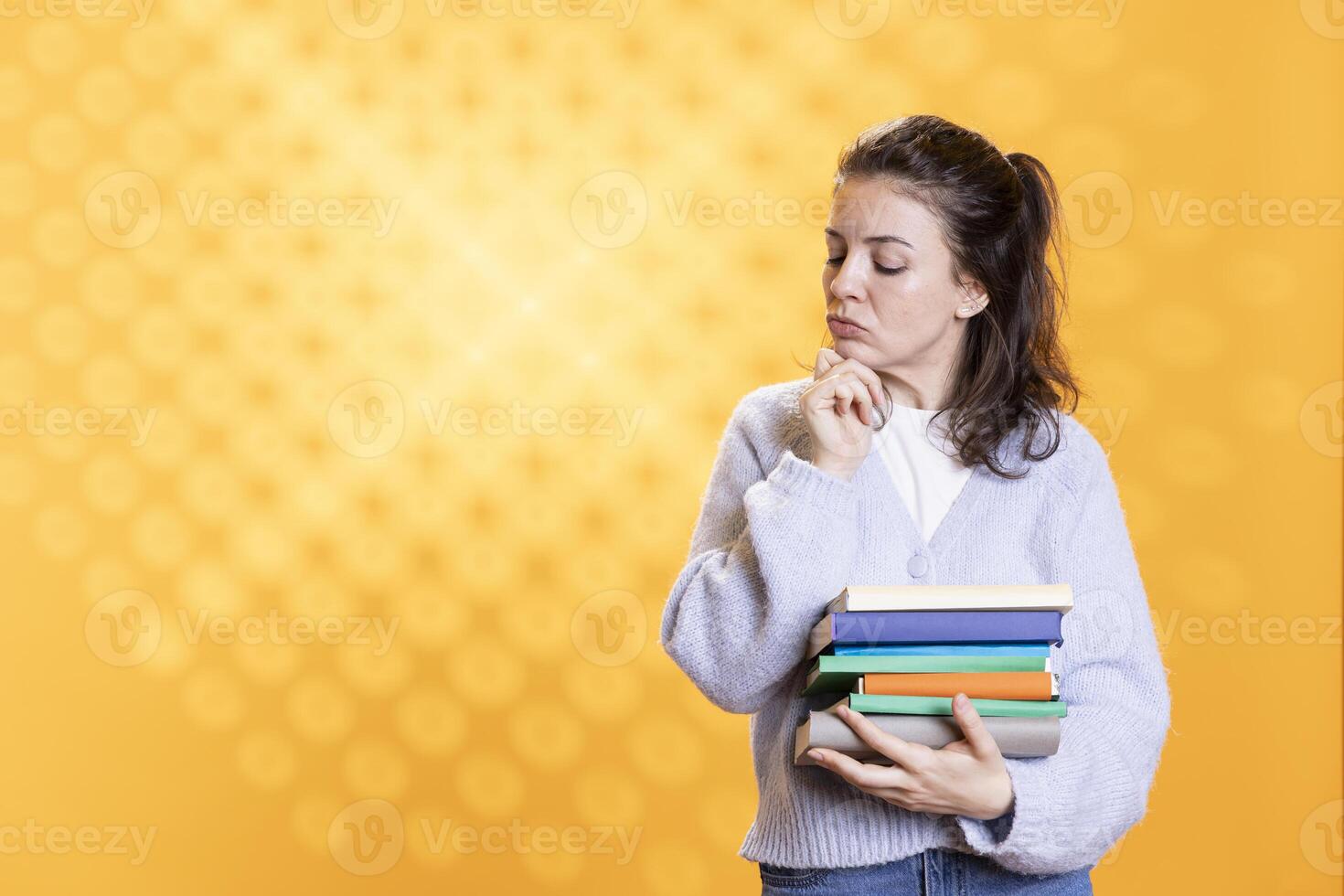 Woman with stack of books in hands deep in thoughts, isolated over studio background. Bookworm holding pile of novels rummaging, thinking about themes and characters after reading photo