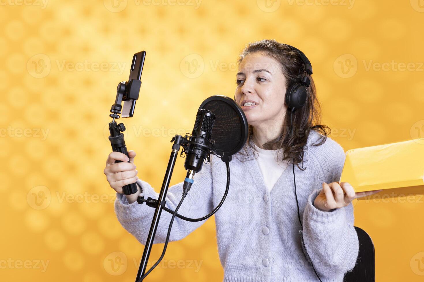 Jolly influencer films vlog at work with selfie stick, reading book to record audiobook. Joyous person doing content creation with mobile phone, recording novel, isolated over yellow studio background photo