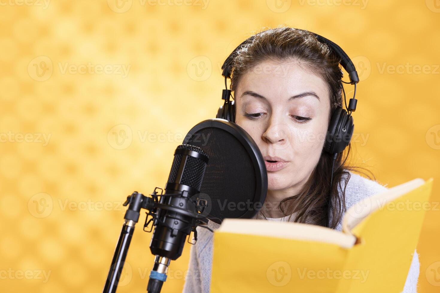 Woman wearing headphones reading aloud from book into microphone against yellow background. Voice actor recording audiobook, creating engaging media content for listeners, doing audio story photo
