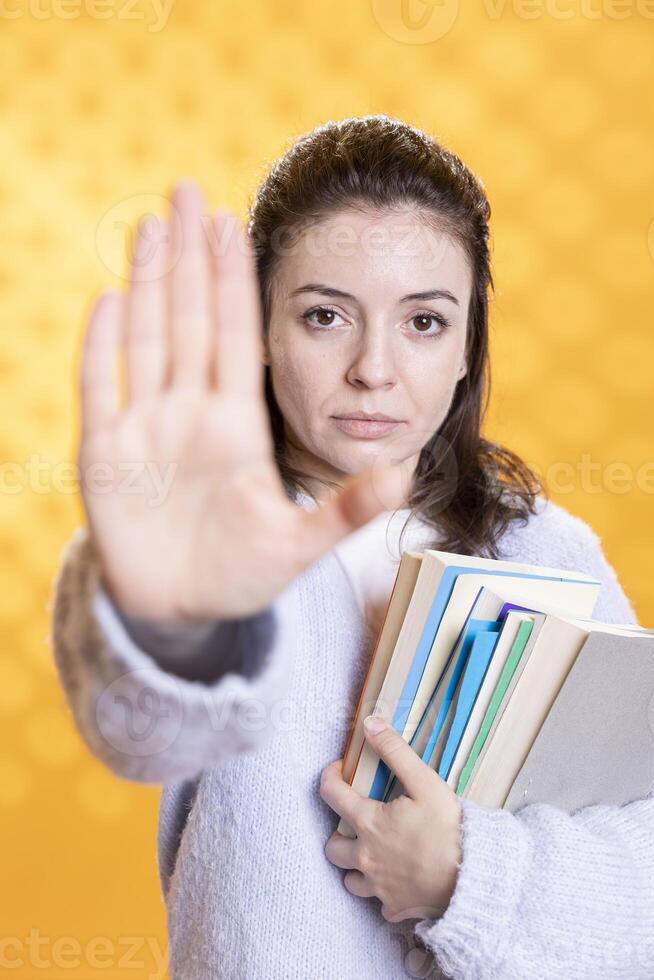 Portrait of annoyed woman doing stop sign while carrying textbooks, tired of preparing for school exam, studio background. Student feeling fed up by university assignments, doing halt hand gesture photo