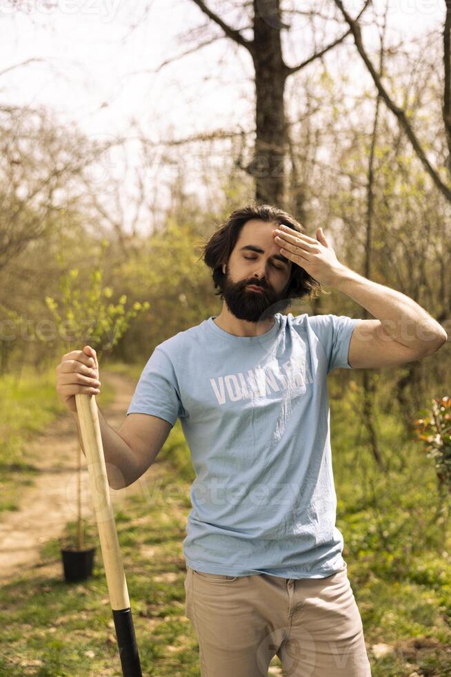 Portrait of overworked exhausted volunteer doing community service, digging holes with a shovel to plant more trees in a forest. Young man activist fighting for reforestation and preservation. photo