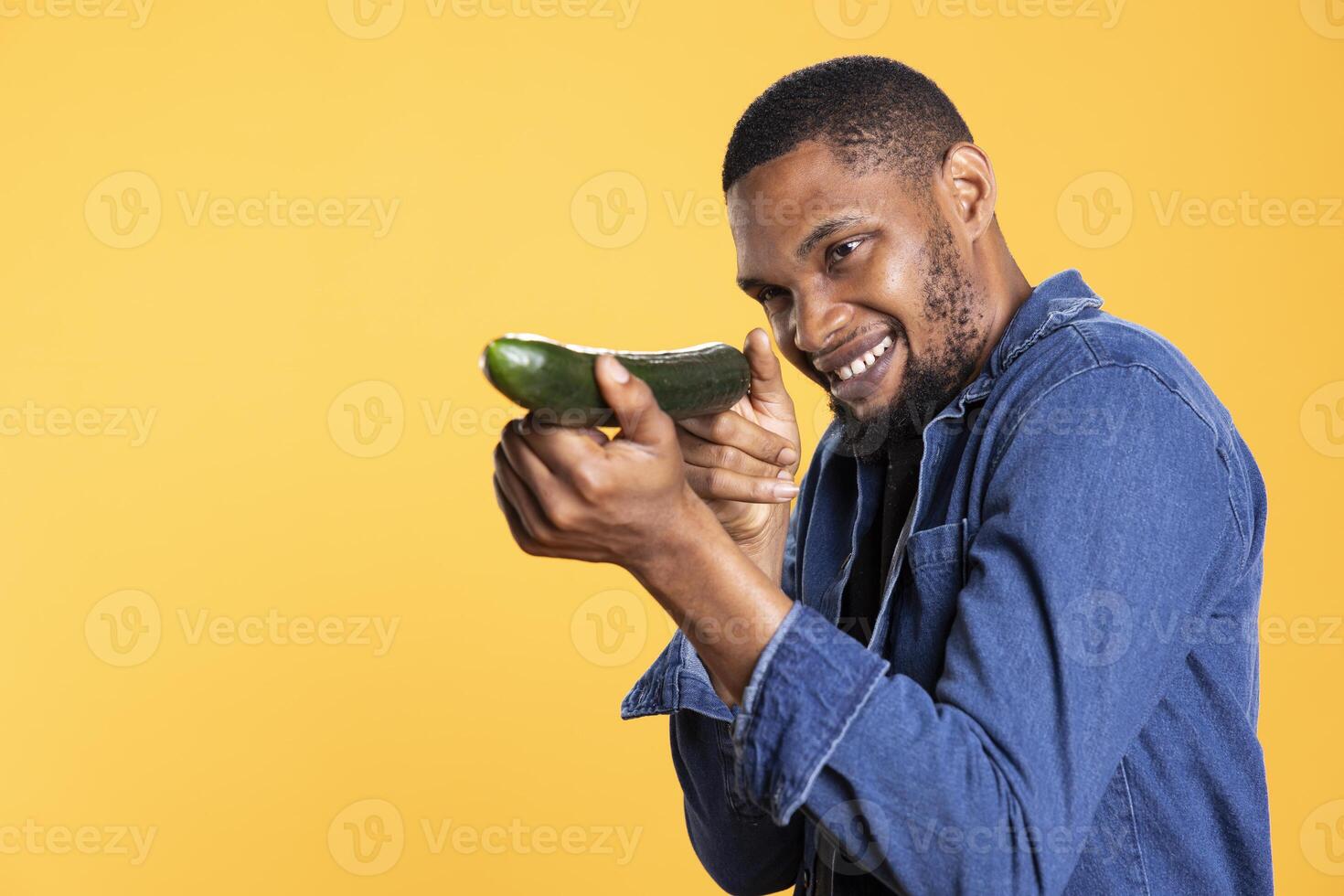 African american funny guy pretending to shoot with a cucumber in studio, playing around and having fun with natural fresh vegetables. Male model acting goofy, posing with raw produce. photo