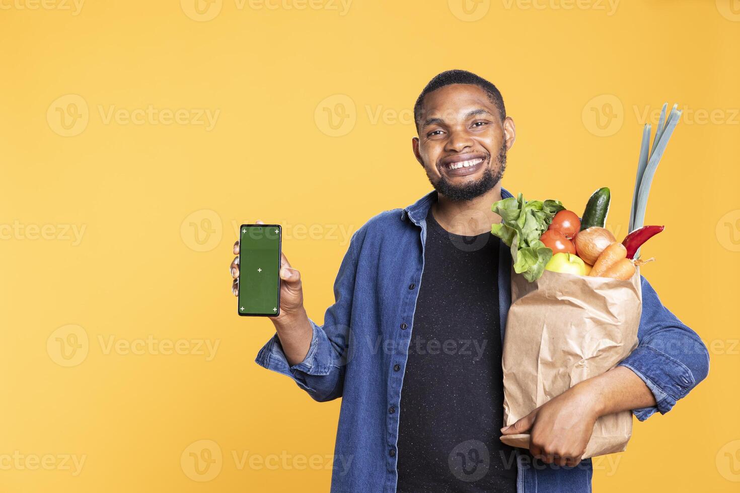 African american guy shows greenscreen display on smartphone, carrying a paper bag with bio produce against yellow background. Male model uses mobile phone with isolated mockup. photo