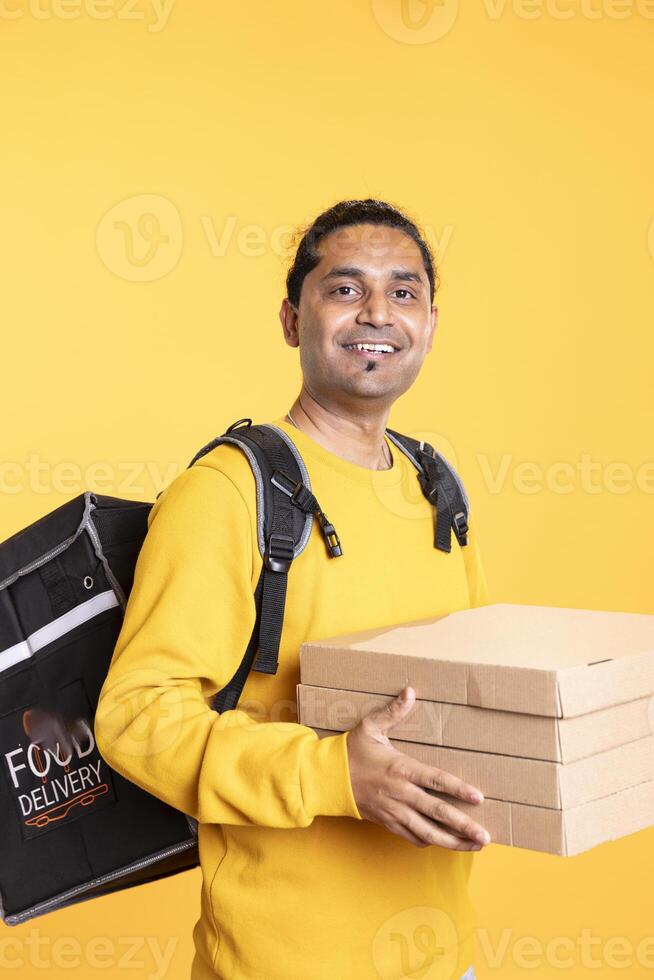 Portrait of happy delivery employee carrying fast food pizza orders, wearing thermal backpack. Upbeat courier holding lunch boxes and transportation bag, rushing to clients, smiling, studio backdrop photo