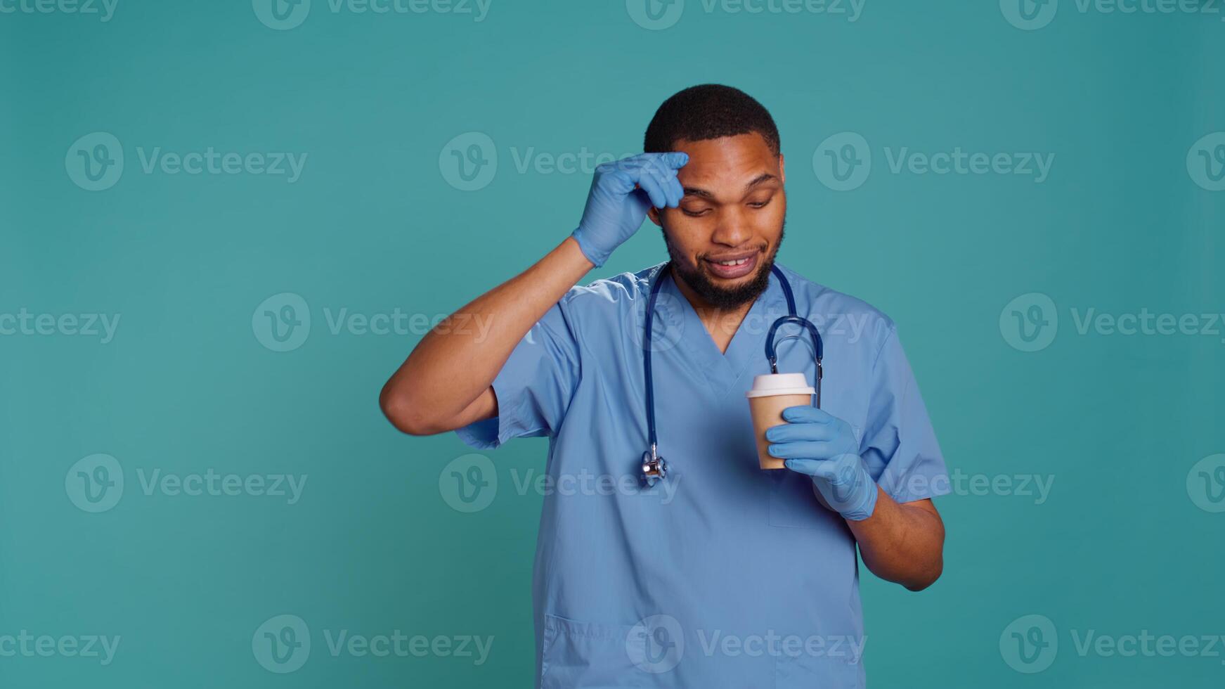 Tired healthcare specialist gaining energy with cup of coffee while at work, enjoying aroma. Fatigued medical practitioner drinking hot beverage, isolated over blue studio background, camera B photo