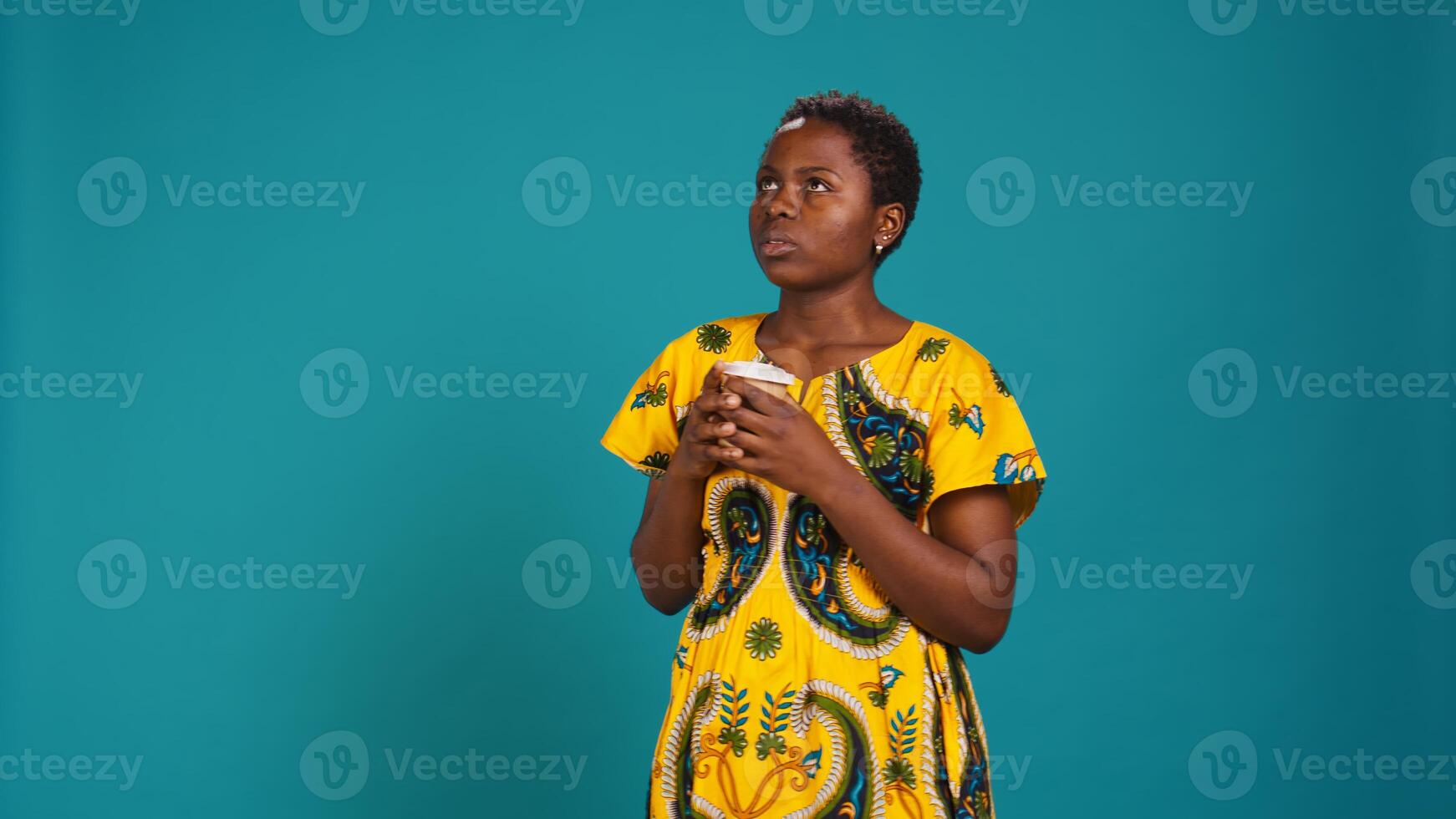Young smiling person enjoying freshness of coffee cup in studio, drinking caffeine refreshment and relaxing against blue background. Happy woman serving a fresh beverage. Camera B. photo